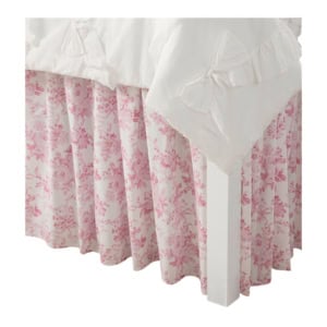 White bedding with bows drapes over the corner of a bed, revealing a pink floral-patterned bed skirt underneath.