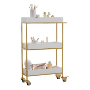 A three-tier white and gold rolling cart holds makeup brushes, cosmetics, and beauty accessories on its shelves.