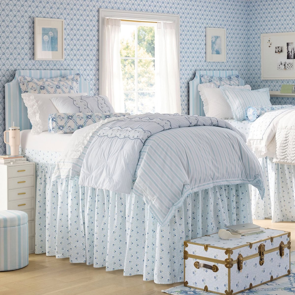 A bedroom with two twin beds in blue and white bedding, floral wallpaper, a window with sheer curtains, and a vintage trunk at the foot of one bed.