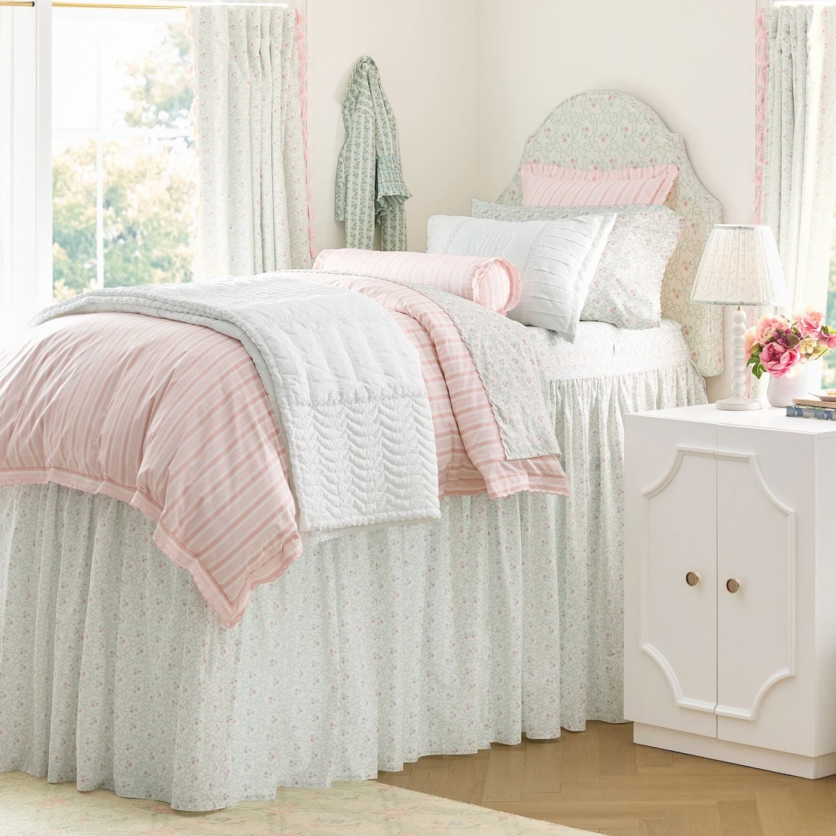 A neatly made bed with pastel pink and white bedding, floral patterns, and ruffled details stands beside a white nightstand with a lamp and flowers in a bright, airy bedroom.