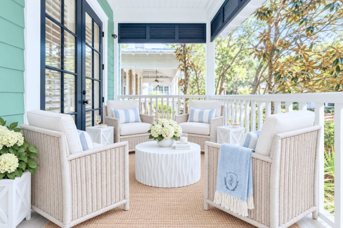 Four white wicker armchairs arranged around a round white coffee table on a covered porch.