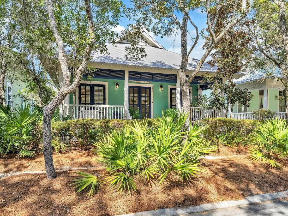 A teal one-story house with a front porch, dark window shutters, and a metal roof, framed by trees and palmettos.