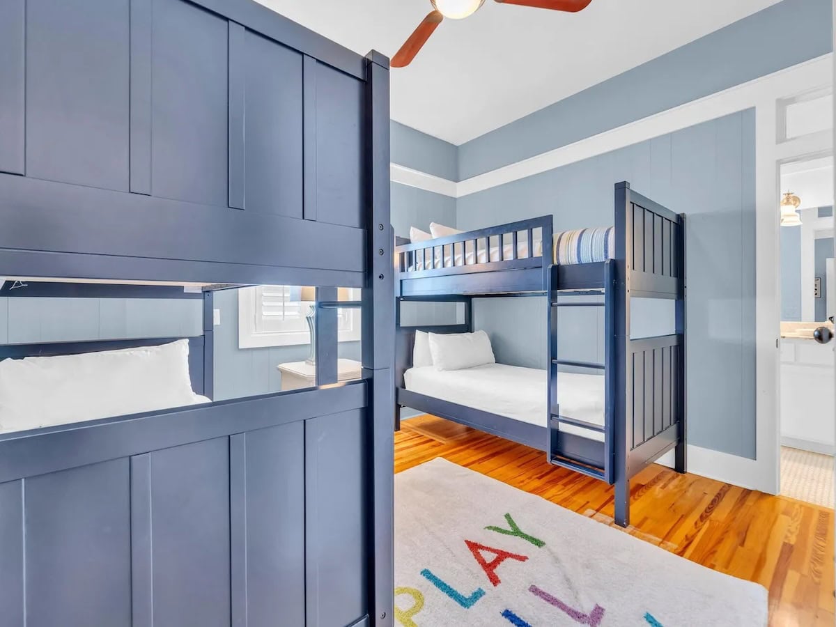 Two navy blue bunk beds in a room with light blue walls, hardwood floors, and a rug with colorful letters.
