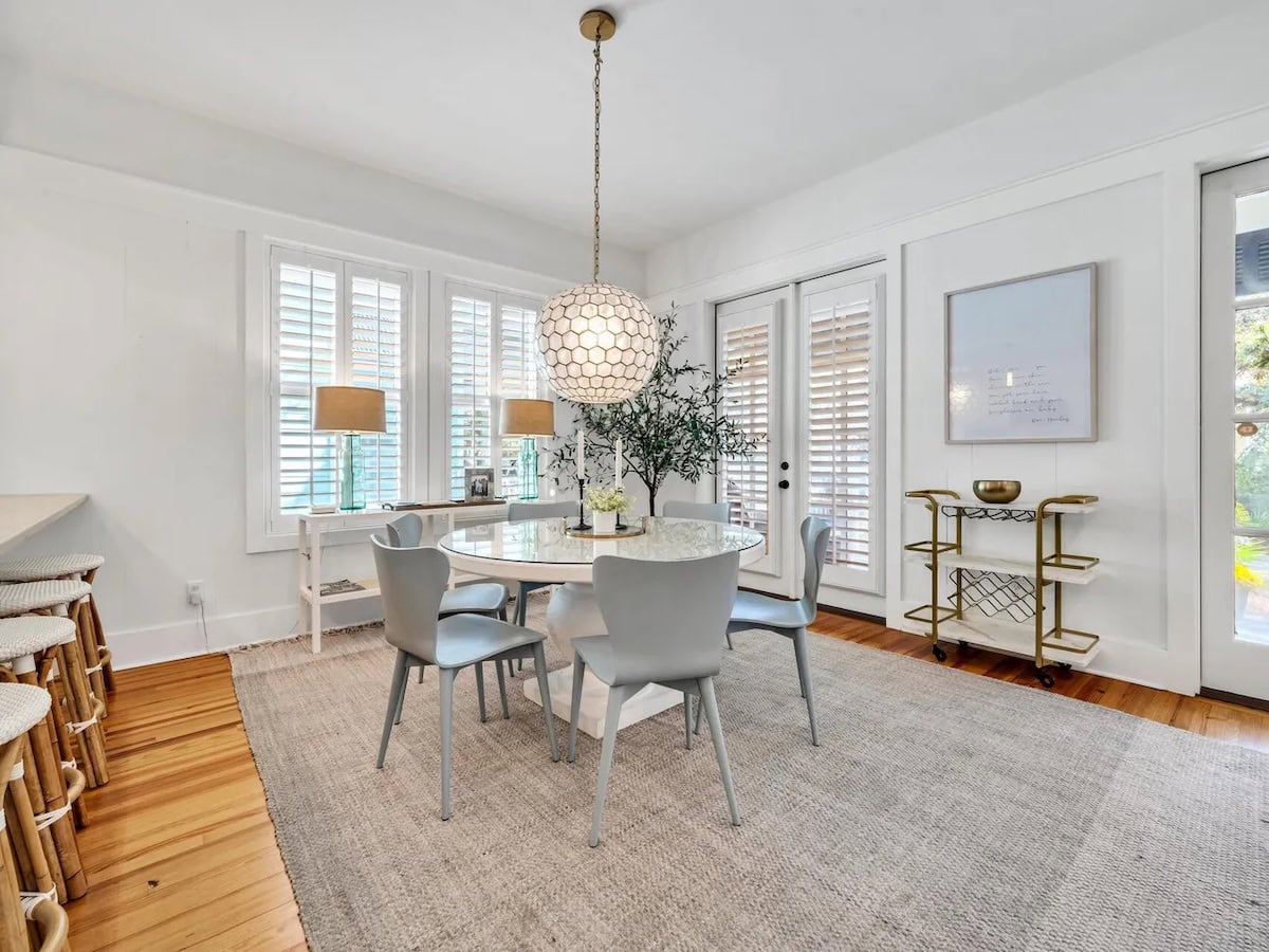 Dining area with a round marble-topped table, four gray chairs, a geometric pendant light, and a bar cart on a large area rug.