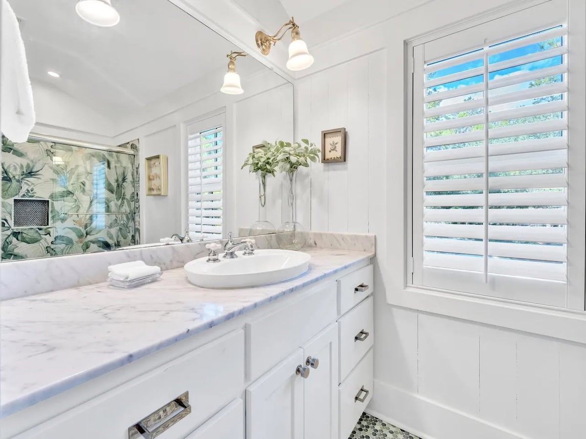 A bathroom vanity with a white marble countertop, oval sink, and white cabinets, featuring a large mirror and window with plantation shutters.