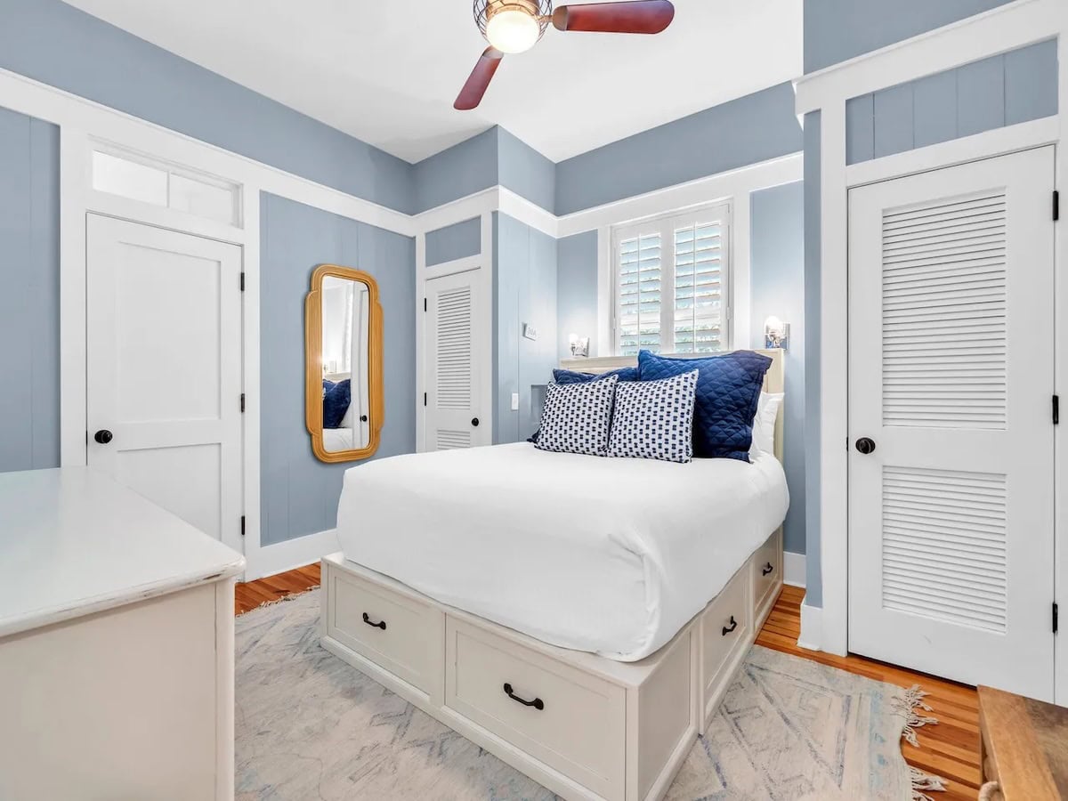 A bedroom with blue walls, a white bed with built-in storage drawers, a wooden-framed mirror, and two white doors.