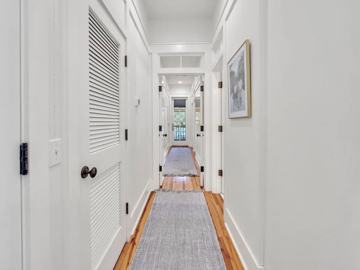 A bright hallway with white paneled walls, a louvered door on the left, and a gray runner on the hardwood floor leading toward a doorway at the end.