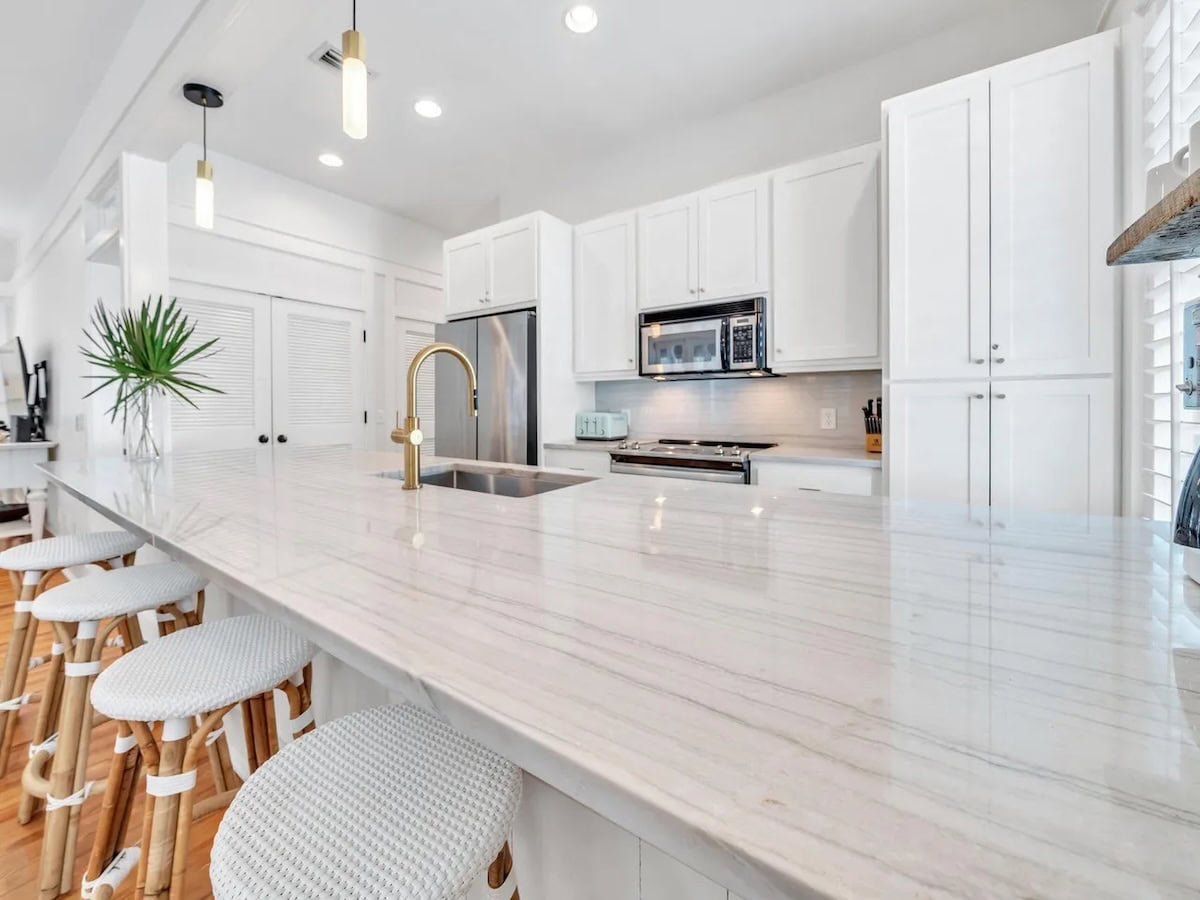 Modern white kitchen with a long marble-topped island, brass faucet, and four cushioned bar stools.