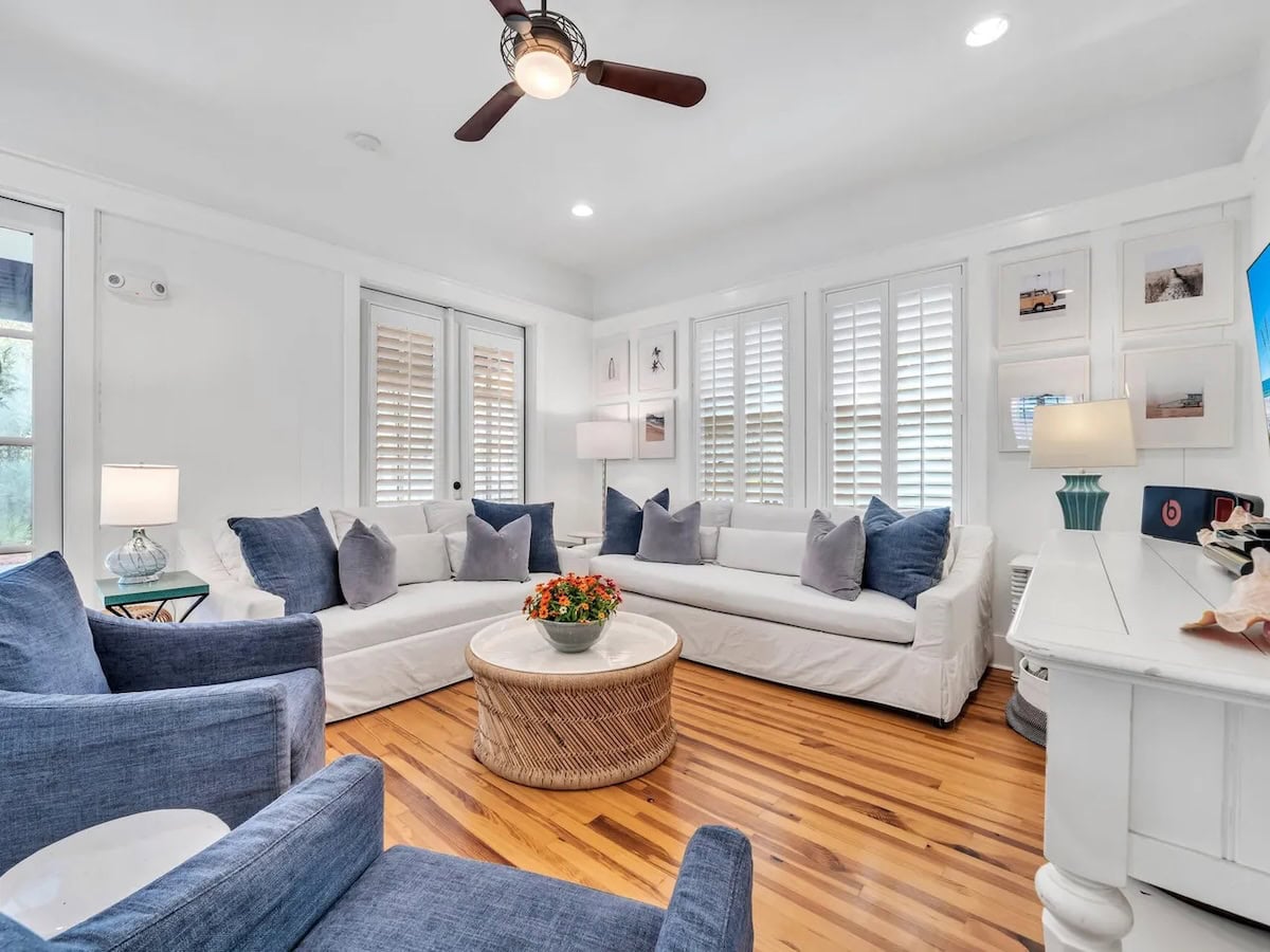 A living room with white slipcovered sofas, two blue armchairs, a round wicker coffee table, and hardwood floors.