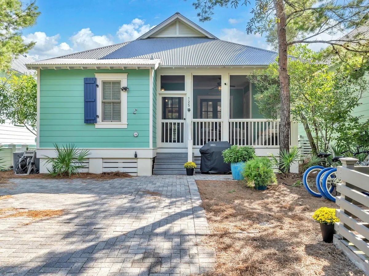 A one-story mint green cottage with a screened-in front porch, a gray metal roof, and a paved driveway.