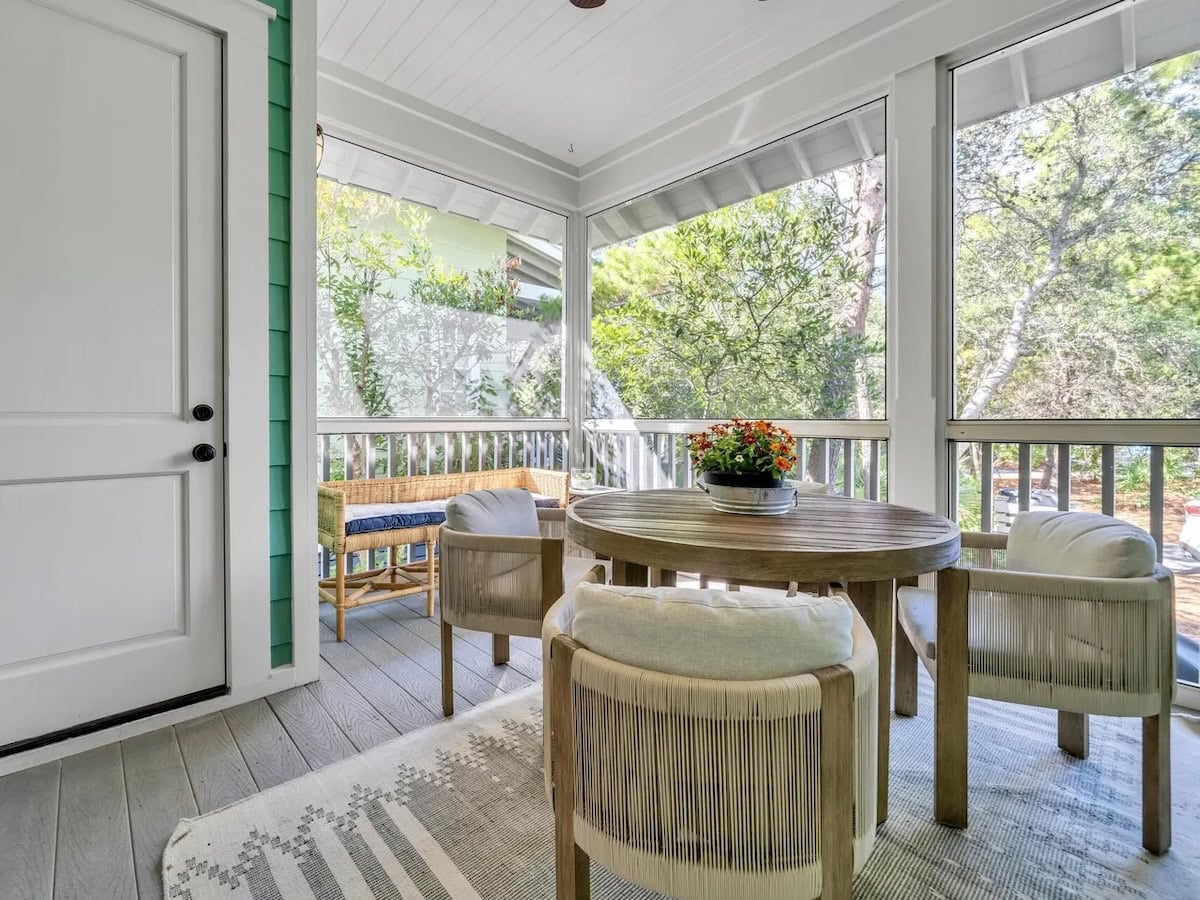 A screened-in porch with a round wooden table, four cushioned chairs, and a small bench, overlooking a wooded area.