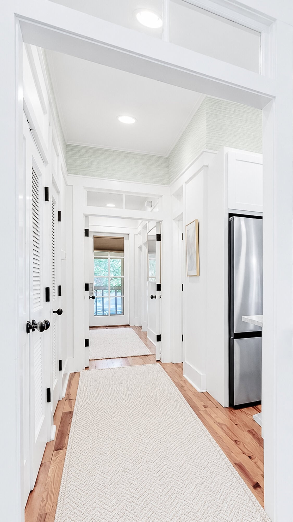 A bright hallway with white walls and wood floors, featuring a long runner rug leading toward a glass-paneled door.