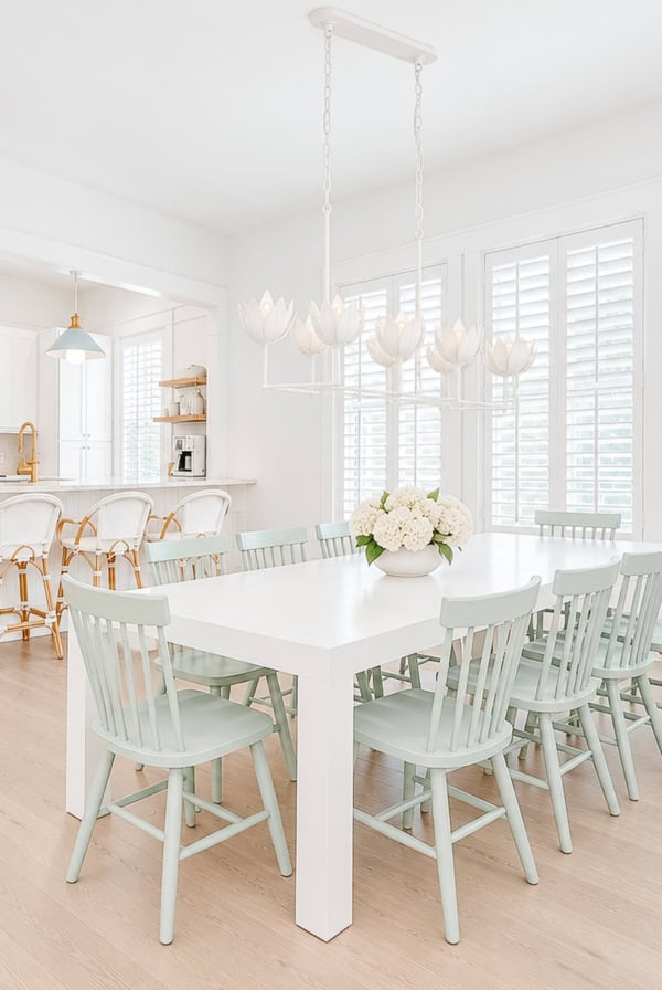 A white dining table with light green chairs and a flower-shaped chandelier in a bright, white-toned room.