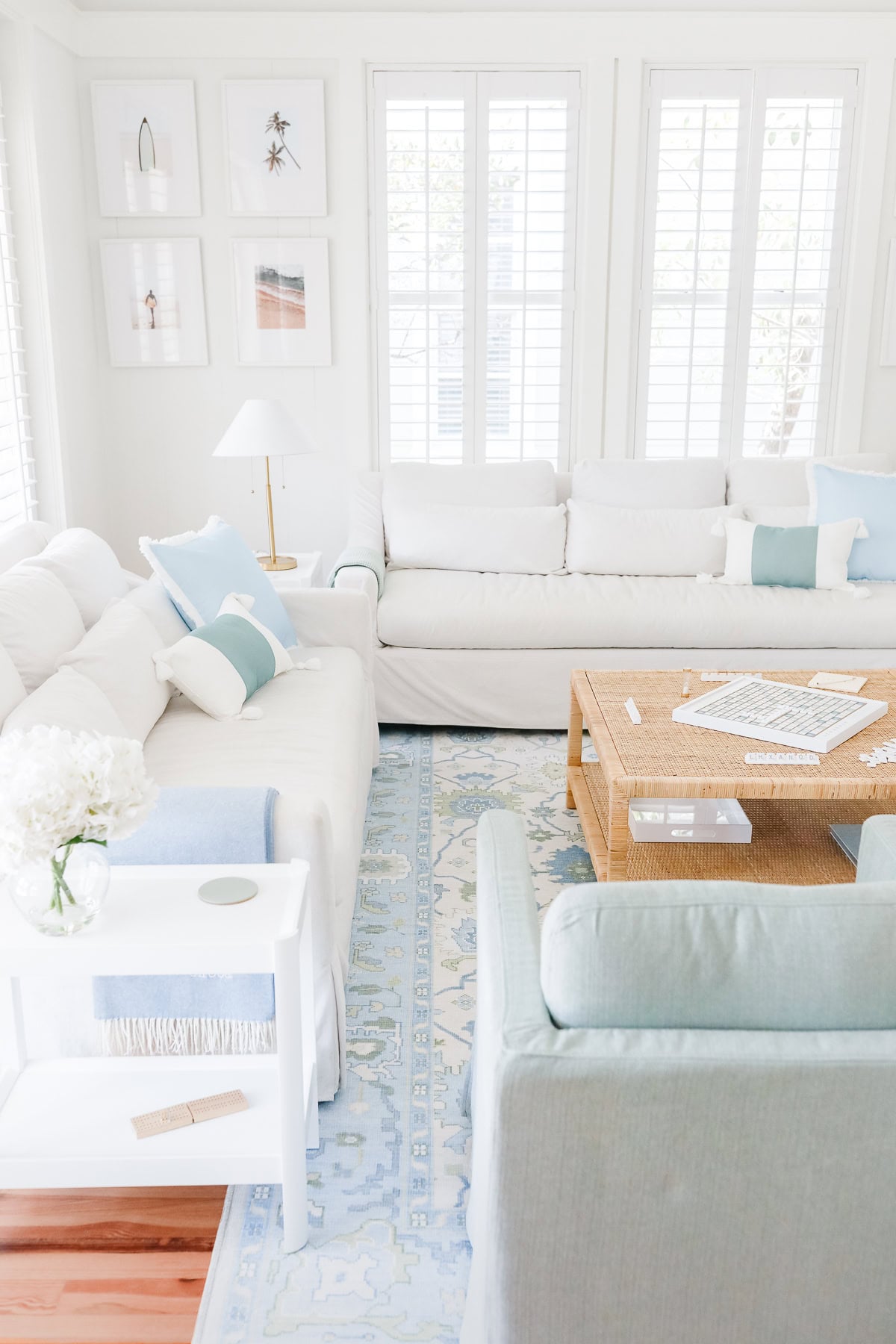 Living room with white sofas, a light blue armchair, a woven coffee table with a game of Scrabble, and a blue and white patterned rug.