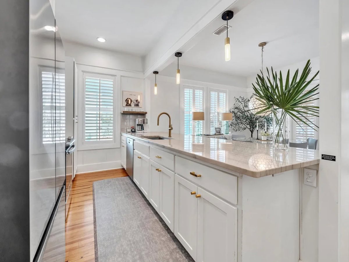 White kitchen with a large island, stone countertops, gold hardware, and a palm leaf centerpiece.