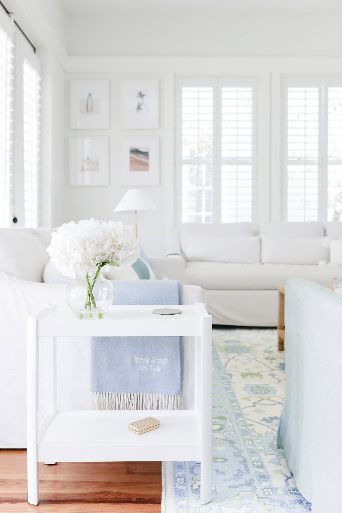 White side table holding a glass vase of hydrangeas and a small object, with a blue blanket reading “Steps Away On 30a” draped over a white sofa in a living room.