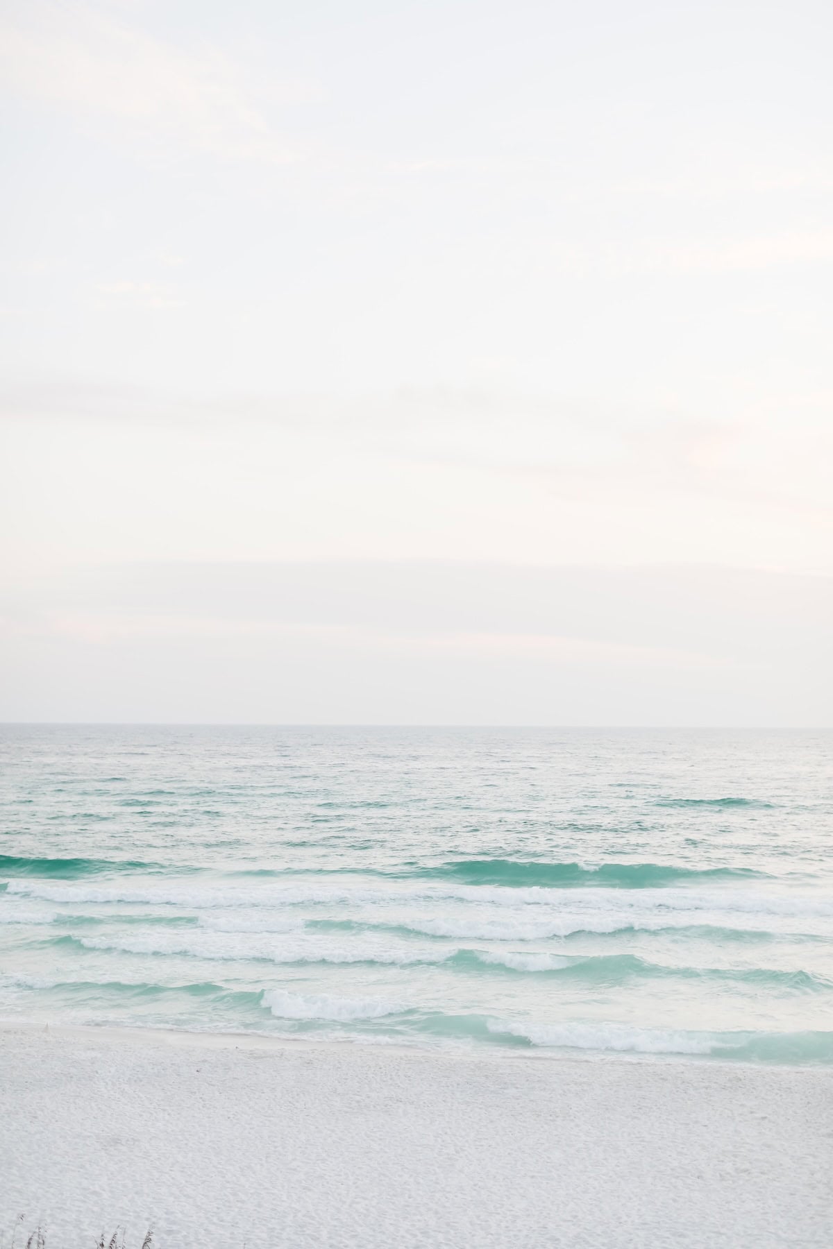 Gentle waves rolling onto a white sand beach under a pale, overcast sky.