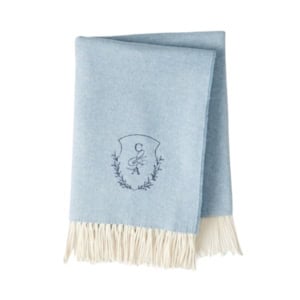 Light blue folded throw blanket with a fringed edge and a monogrammed crest embroidered in the center.