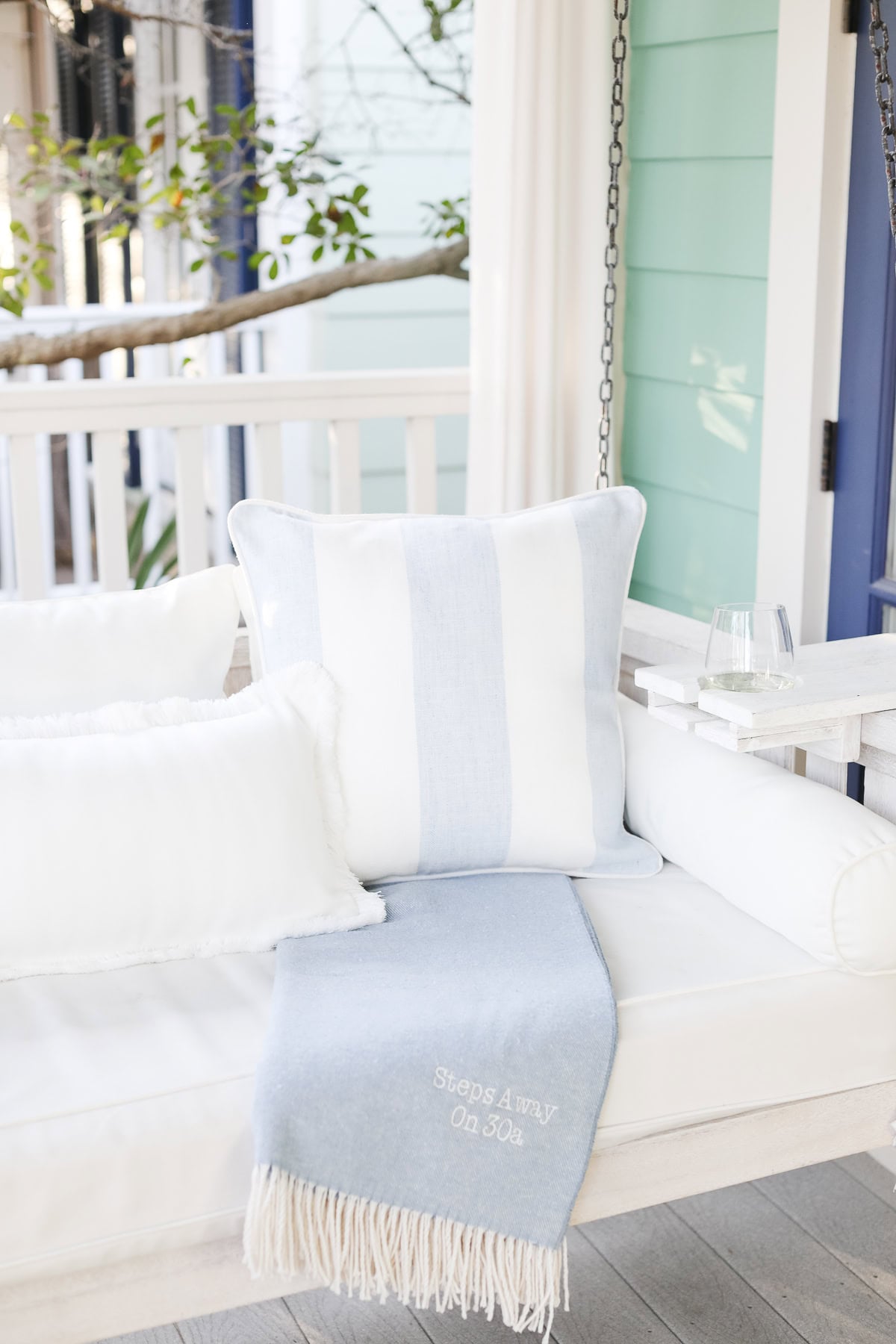 A white porch swing with white pillows, a blue and white striped pillow, and a light blue throw blanket embroidered with “Steps Away On 30a.”