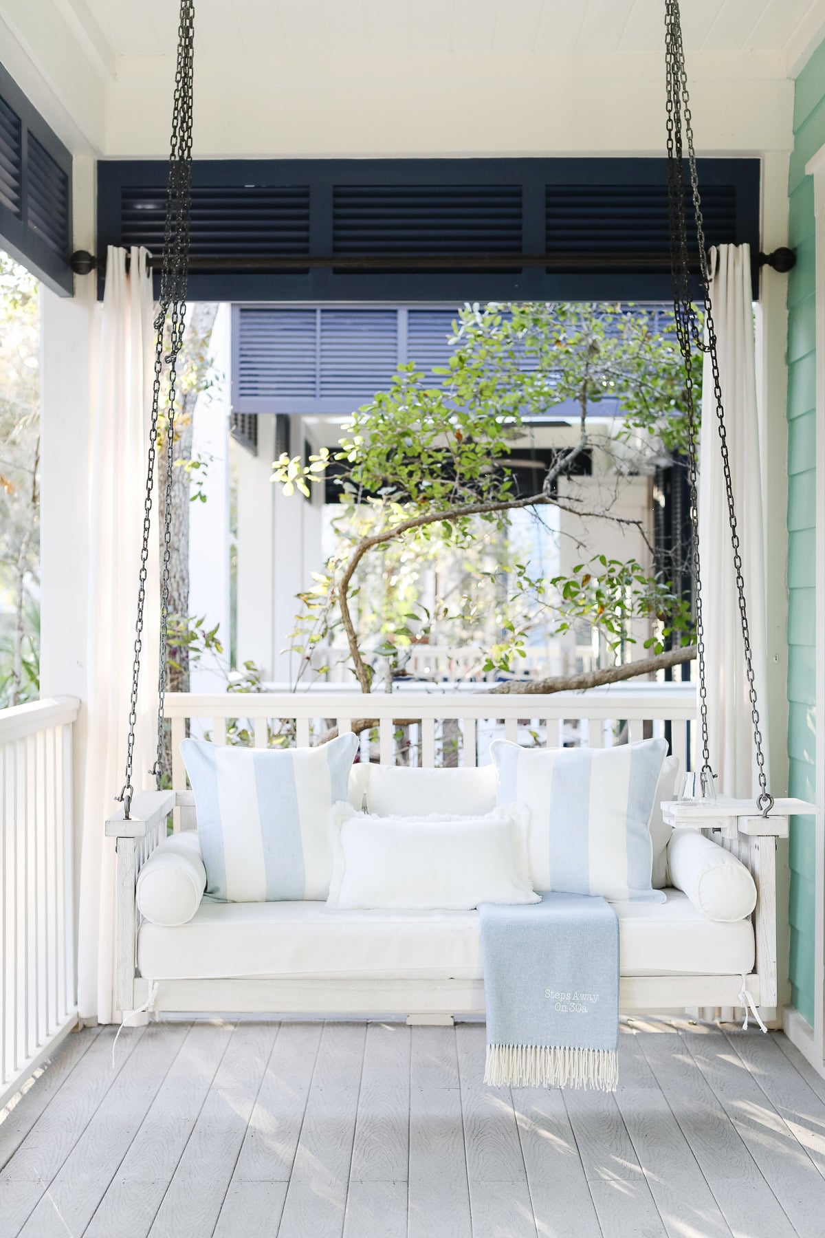 A white wooden porch swing hanging by chains, decorated with blue-and-white striped pillows and a light blue throw blanket.