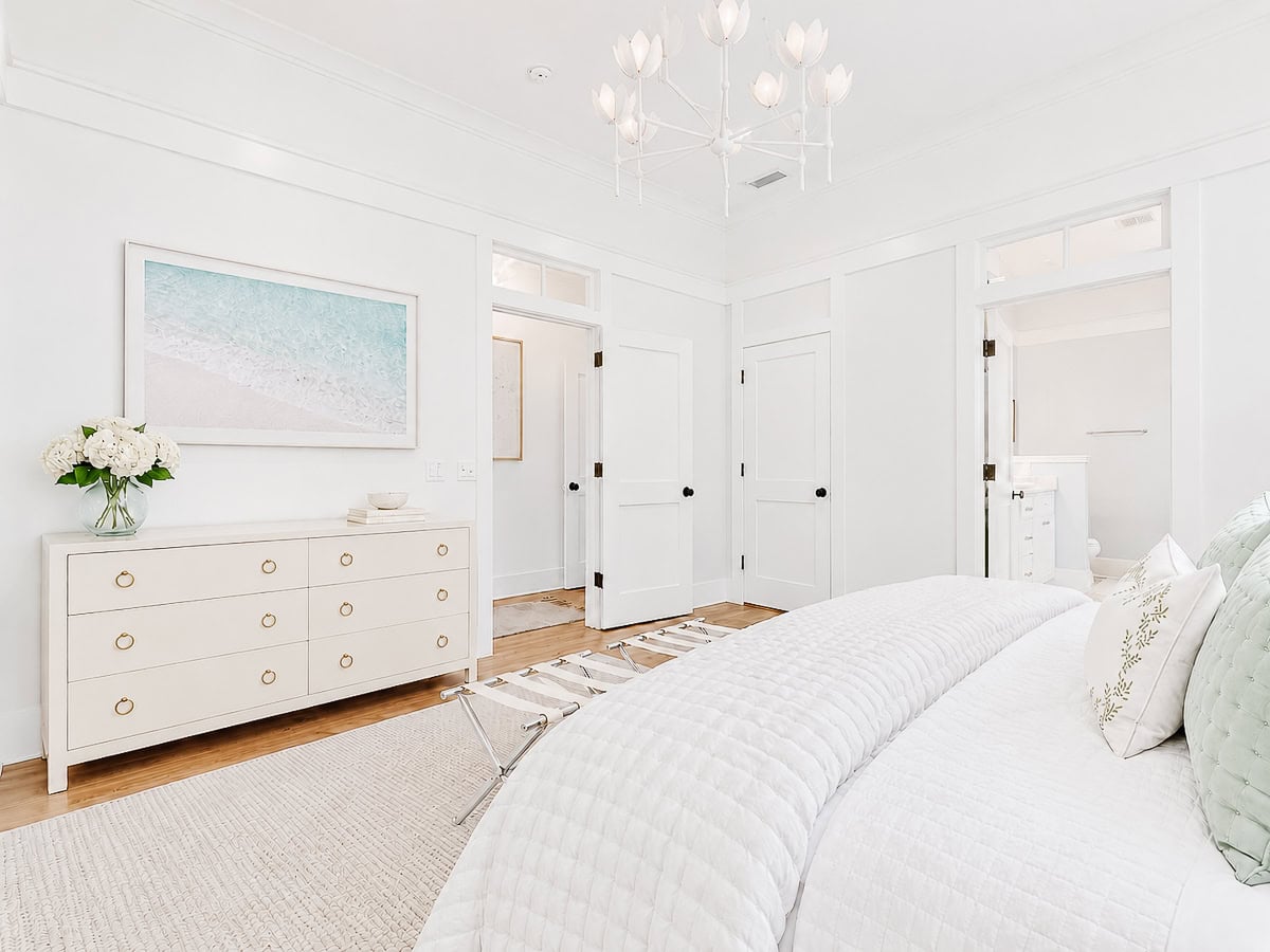 A white bedroom featuring a dresser with a vase of hydrangeas, a framed beach photograph, and a bed with quilted white bedding.