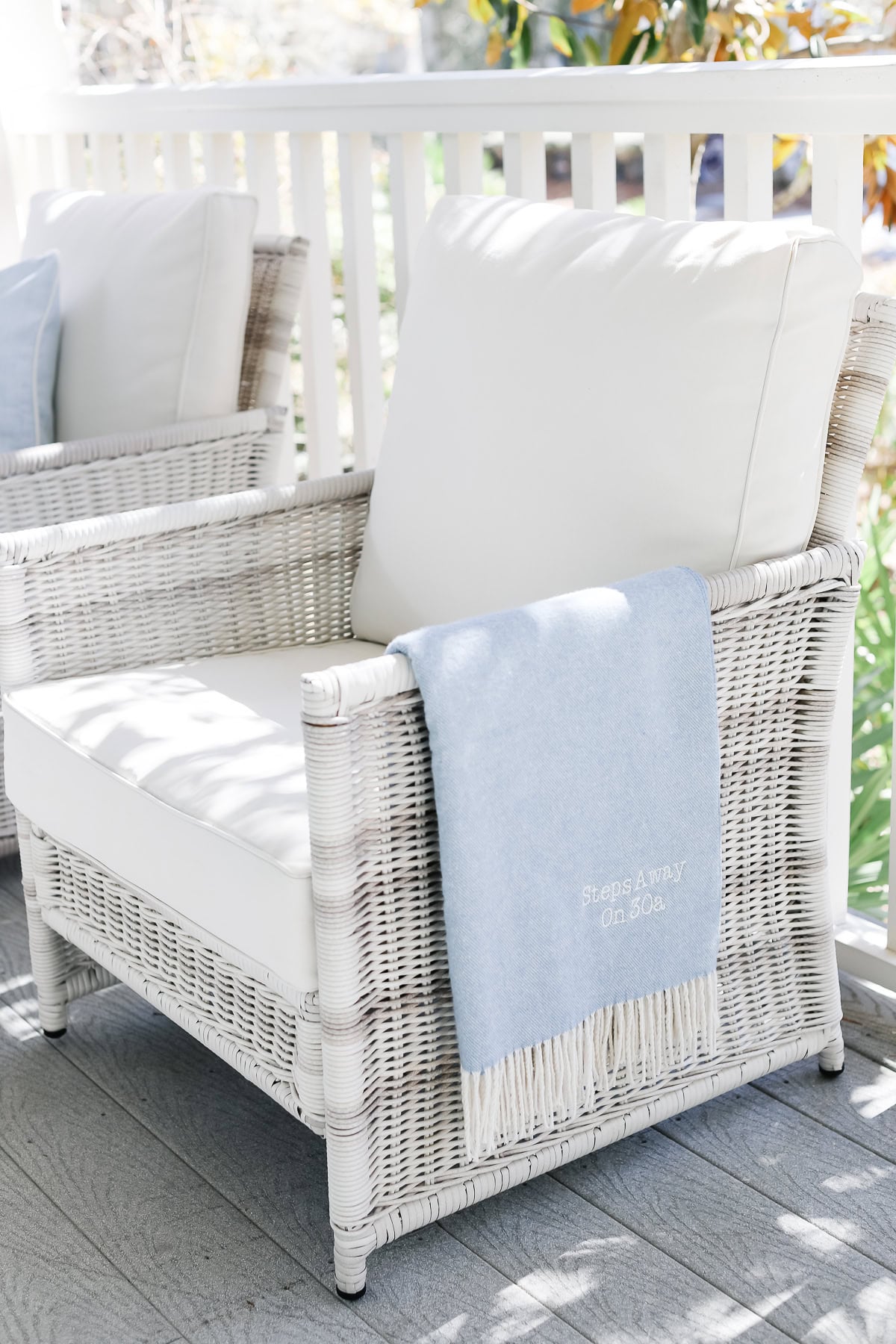White wicker outdoor chair with white cushions and a light blue throw blanket featuring the embroidered text “Steps Away On 30A.”