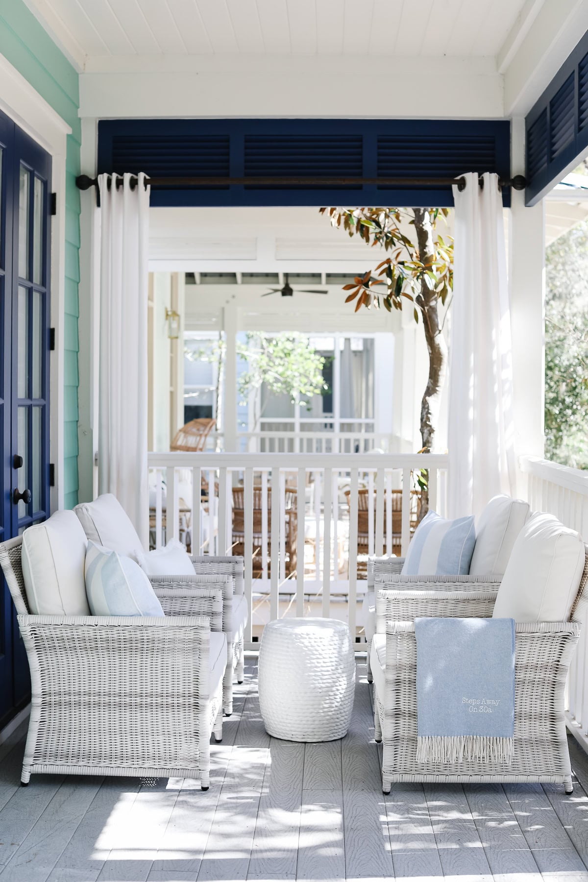 Two white wicker armchairs with cushions and a white drum stool on a covered porch.