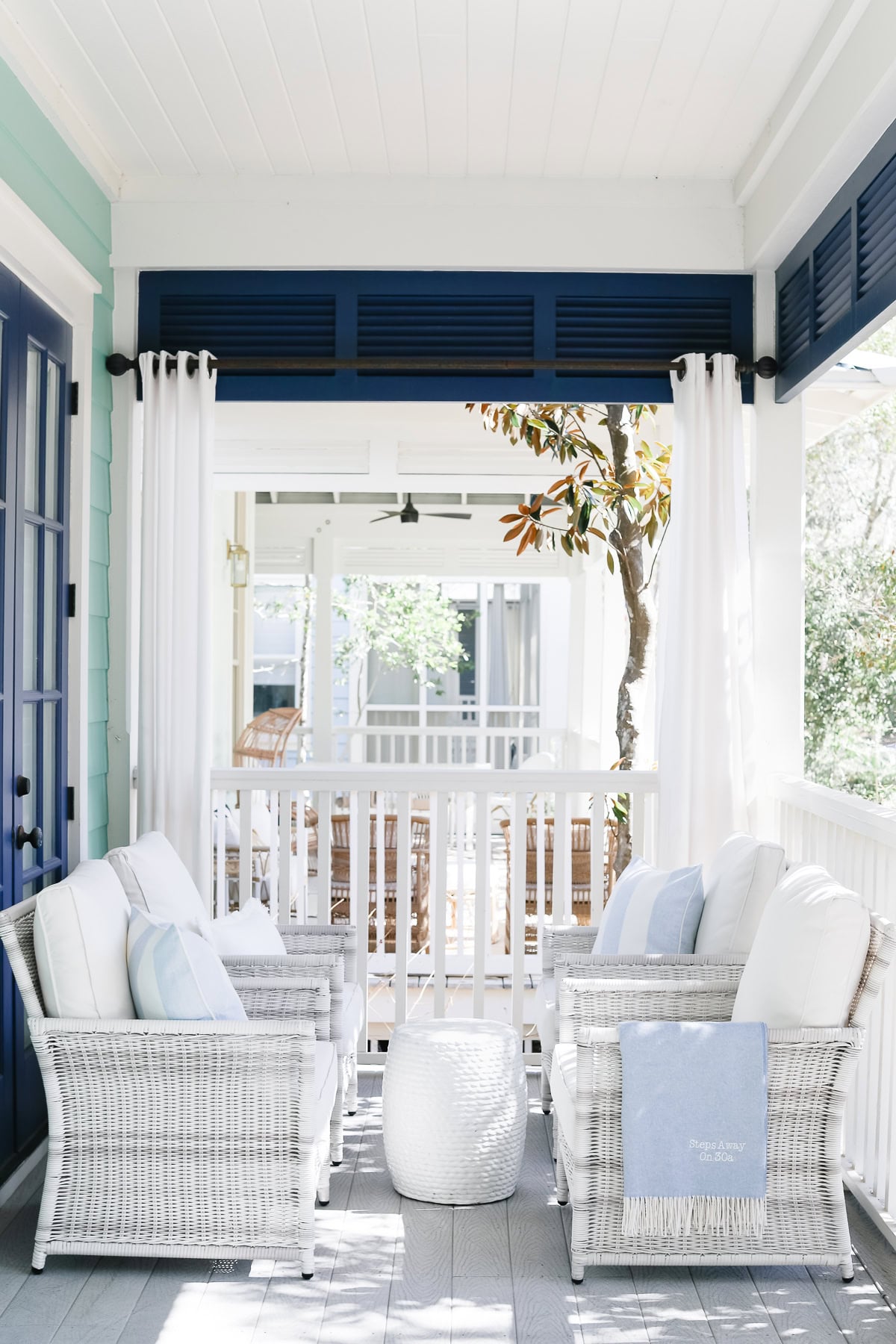 A covered porch with two white wicker armchairs facing each other and a small white stool in between, with white curtains and a view of another porch across a railing.